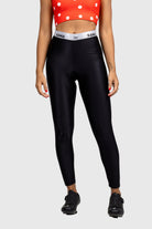 A pair of women's pants with a black color scheme, featuring a high waist and a branded elastic band at the waist. The pants are paired with a dotted orange top and black sports bra visible in the background.